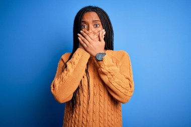 Genç Afro-Amerikalı kadın, günlük sarı süveter giyip mavi izole arka planda dikiliyor. Ağzını yanlışlıkla ellerle kapatıyor. Gizli konsept.