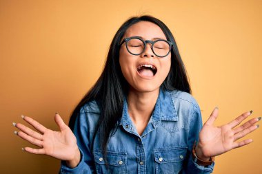 İzole edilmiş sarı arka plan üzerine sıradan kot gömlek giyen genç, güzel Çinli bir kadın başarı için deli ve çılgınlığı kollarını kaldırarak ve gözlerini kapayarak heyecanla bağırarak kutluyor. Kazanan konsept