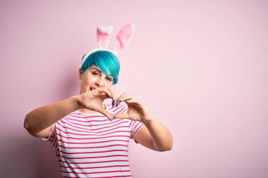 Young woman with fashion blue hair wearing easter rabbit ears over pink background smiling in love doing heart symbol shape with hands. Romantic concept.