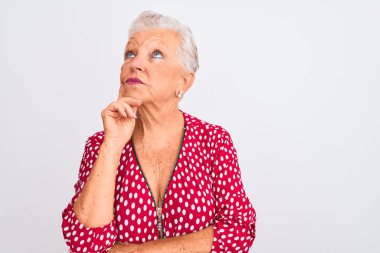 Senior grey-haired woman wearing red casual jacket standing over isolated white background with hand on chin thinking about question, pensive expression. Smiling with thoughtful face. Doubt concept.