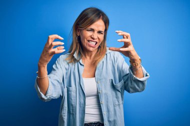 Middle age beautiful woman wearing casual shirt standing over isolated blue background Shouting frustrated with rage, hands trying to strangle, yelling mad