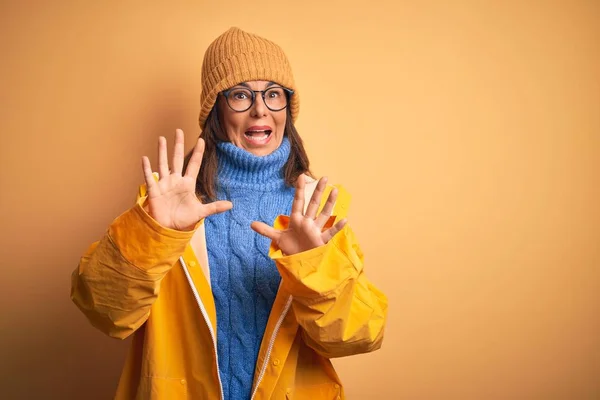 Middle age woman wearing yellow raincoat and winter hat over isolated background afraid and terrified with fear expression stop gesture with hands, shouting in shock. Panic concept.
