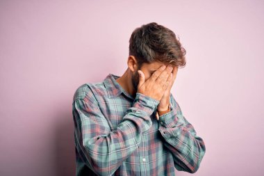 Young handsome man with beard wearing casual shirt standing over pink background with sad expression covering face with hands while crying. Depression concept.