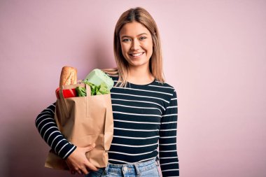 Genç, güzel bir kadın, elinde kağıt torbayla pembe bir arka plandan alışveriş yapıyor. Mutlu bir yüzü var, gülümsüyor ve kendine güvenen bir gülümsemeyle dişlerini gösteriyor.