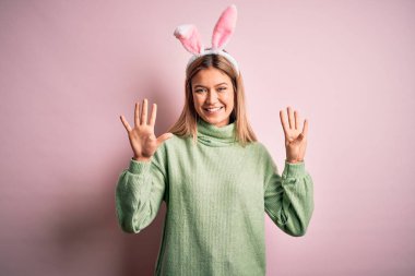 Paskalya tavşanı kulakları takan genç ve güzel bir kadın pembe arka planda dikilip dokuz numaralı parmaklarıyla işaret ederken kendinden emin ve mutlu bir şekilde gülümsüyor..