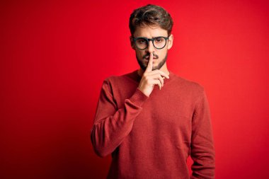 Young handsome man with beard wearing glasses and sweater standing over red background asking to be quiet with finger on lips. Silence and secret concept.