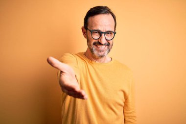 Orta yaşlı, beyaz bir adam, günlük süveter ve gözlük takıyor, sarı arka planda mutlu bir şekilde gülümsüyor, yardım ve kabullenme eli uzatıyor..