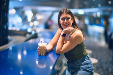 Mutlu ve kendinden emin gülümseyen genç ve güzel bir kadın. Lokantada elinde bir bardak kahveyle tezgaha yaslanıyorsun.