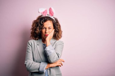 Orta yaşlı, tavşan kulaklı güzel bir kadın pembe arka planda dikilip kollarını kavuşturarak depresyondan sıkılmış ve yorgun göründüğünü düşünüyor..