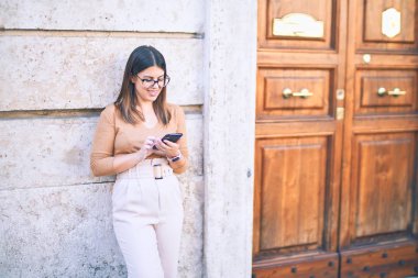 Mutlu ve kendinden emin gülümseyen genç ve güzel bir kadın. Yüzünde gülümseme, duvarlara yaslanmış bir şekilde şehirde akıllı telefon kullanarak dikiliyorsun.