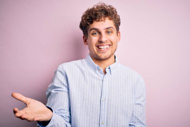 Sarışın, kıvırcık saçlı, beyaz arka planda çizgili tişört giyen, neşeli, kollarını açıp gülümseyen, arkadaş canlısı, olumlu ve kendine güvenen genç bir adam.