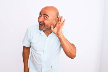 Middle age handsome man wearing casual shirt standing over isolated white background smiling with hand over ear listening an hearing to rumor or gossip. Deafness concept.
