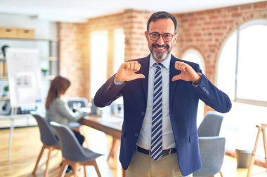 Middle age handsome businessman wearing glasses   standing at the office looking confident with smile on face, pointing oneself with fingers proud and happy. clipart