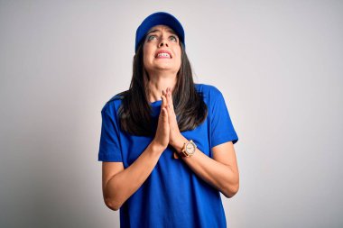 Mavi arka planda duran mavi şapkalı genç teslimatçı kadın elleri bağlı yalvarıyor ve dua ediyor. Yüzünde umut ifadesi var. Çok duygusal ve endişeli. Yalvarıyorum..