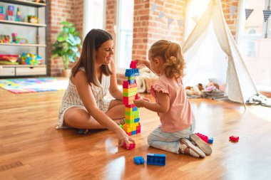 Güzel öğretmen ve sarışın küçük kız kreşteki plastik blokları kullanarak kule inşa ediyorlar.