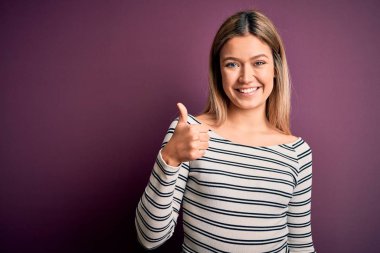 Genç, güzel, sarışın bir kadın, mor izole edilmiş arka plan üzerine sıradan çizgili tişört giyip el hareketleriyle mutlu mesut hareket ediyor. Kameraya bakıp başarıyı gösteren ifade onaylanıyor.