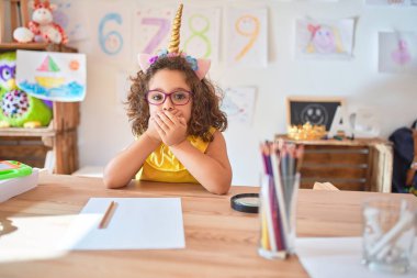 Beautiful toddler wearing glasses and unicorn diadem sitting on desk at kindergarten shocked covering mouth with hands for mistake. Secret concept.