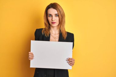 Redhead business caucasian woman holding banner over yellow isolated background with a confident expression on smart face thinking serious clipart