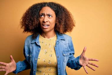 Afro saçlı, sarı arka plan üzerine günlük kot gömlek giyen, deli gibi bağıran, agresif ifade ve kollarıyla bağıran Afro-Amerikalı genç bir kadın. Hayal kırıklığı konsepti.