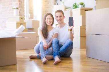 Beautiful couple sitting on the floor holding smartphone at new home around cardboard boxes with a happy face standing and smiling with a confident smile showing teeth