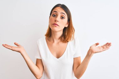 Beautiful redhead woman wearing casual white t-shirt over isolated background clueless and confused expression with arms and hands raised. Doubt concept.