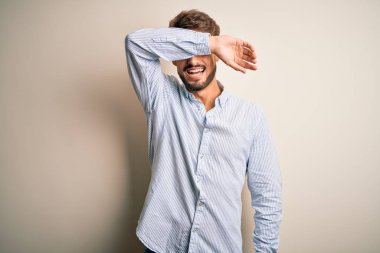 Young handsome man with beard wearing striped shirt standing over white background covering eyes with arm smiling cheerful and funny. Blind concept.