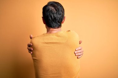 Orta yaşlı, ağarmış, günlük süveter ve gözlük takan, izole edilmiş sarı arka planda kendine sarılan mutlu ve pozitif bir adam. Sevgi ve özgüven.