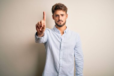 Young handsome man with beard wearing striped shirt standing over white background Pointing with finger up and angry expression, showing no gesture