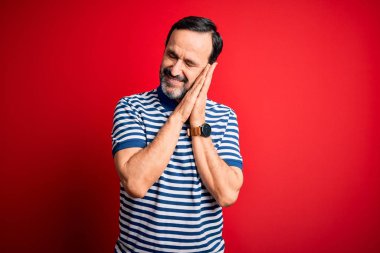 Orta yaşlı, beyaz, sıradan çizgili polo giyen, izole edilmiş kırmızı arka planda duran, yorgun, hayal kuran, elleri ve gözleri kapalı gülümseyerek poz veren bir adam..