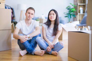 Young beautiful couple sitting on the floor at new home around cardboard boxes with serious expression on face. Simple and natural looking at the camera.