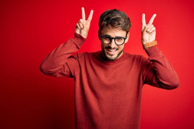 Young handsome man with beard wearing glasses and sweater standing over red background Posing funny and crazy with fingers on head as bunny ears, smiling cheerful