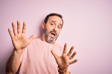 Orta yaşlı, beyaz tişörtlü bir adam pembe arka plan üzerinde dikiliyor korku ifadesinden korkuyor ve dehşete düşüyor. El hareketlerini durdur, şok içinde bağır. Panik konsepti.