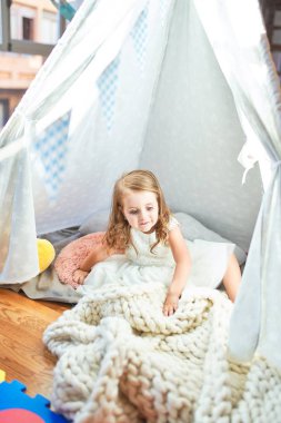 Anaokulunun içinde battaniyenin üzerinde oturan sevimli sarışın çocuk.