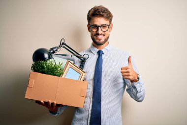 Young businessman with beard fired holding cardboard standing over white background happy with big smile doing ok sign, thumb up with fingers, excellent sign