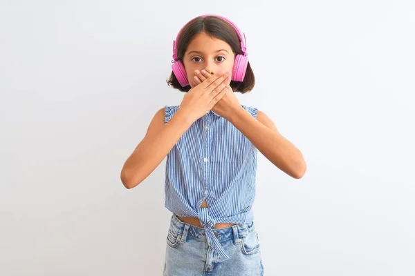 孤立した白い背景の上にヘッドフォンを使用して音楽を聞いて美しい子供の女の子は間違いのための手で口をカバーショックを受けた 秘密概念 — ストック写真