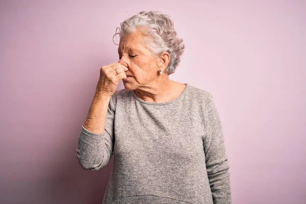 Senior beautiful woman wearing casual t-shirt standing over isolated pink background smelling something stinky and disgusting, intolerable smell, holding breath with fingers on nose. Bad smell