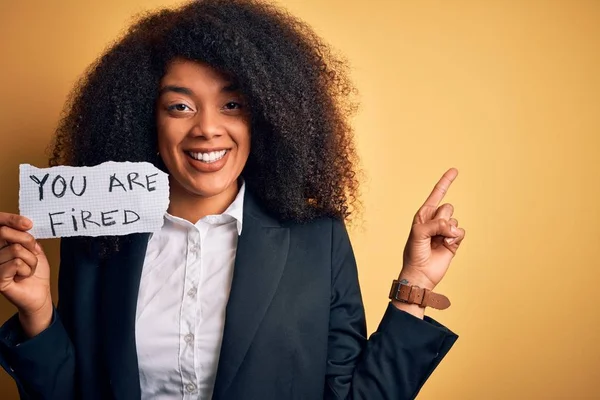 African american business boss woman with afro hair holding you are fired paper for dismissal very happy pointing with hand and finger to the side