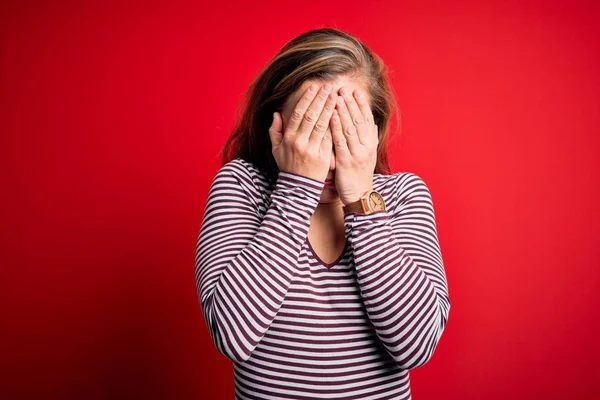 Young beautiful blonde woman wearing casual striped t-shirt over isolated red background with sad expression covering face with hands while crying. Depression concept.