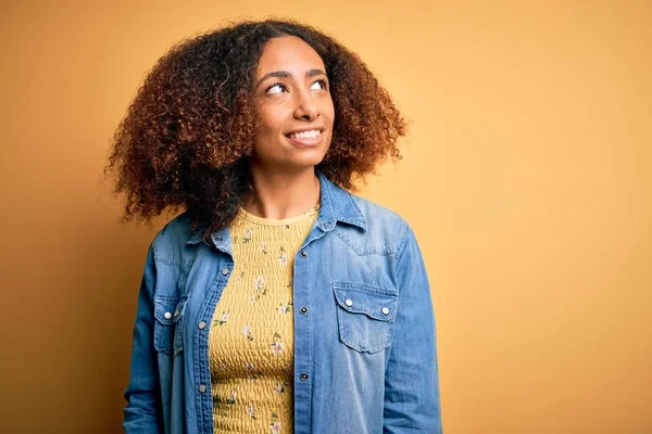 Young african american woman with afro hair wearing casual denim shirt over yellow background looking away to side with smile on face, natural expression. Laughing confident.
