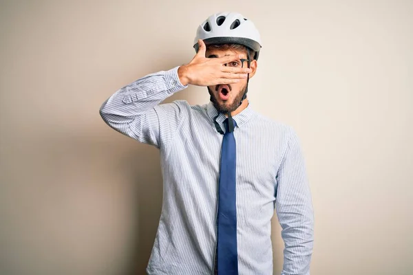 Young businessman wearing glasses and bike helmet standing over isolated white bakground peeking in shock covering face and eyes with hand, looking through fingers with embarrassed expression.