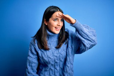 Young brunette woman with blue eyes wearing casual turtleneck sweater very happy and smiling looking far away with hand over head. Searching concept.