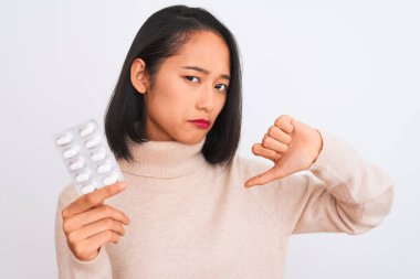 Young beautiful chinese woman holding pills standing over isolated white background with angry face, negative sign showing dislike with thumbs down, rejection concept