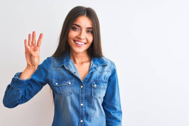 İzole edilmiş beyaz arka plan üzerinde duran sıradan bir kot gömlek giyen genç ve güzel bir kadın dört numaralı parmaklarıyla işaret ederken kendinden emin ve mutlu bir şekilde gülümsüyor..