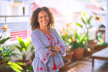 Orta yaşlı güzel bir kadın mutlu ve kendinden emin gülümsüyor terasta yüzünde bir gülümsemeyle duruyor.