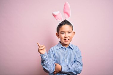 Paskalya tavşanı kulakları takan küçük bir çocuk pembe arka planda, yüzünde kocaman bir gülümseme, eli ve parmağı kameraya bakıyor..