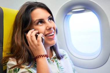 Havaalanında oturan genç yolcu kadın. Pencereden gökyüzü manzaralı. Akıllı telefon kullanarak arıyor.