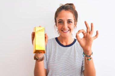 Young beautiful redhead woman holding gold ingot over isolated background doing ok sign with fingers, excellent symbol