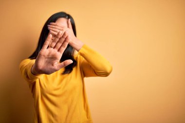 Young brunette woman with blue eyes wearing casual sweater over yellow background covering eyes with hands and doing stop gesture with sad and fear expression. Embarrassed and negative concept.