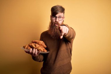 Redhead Irish man with beard eating french croissant pastry over yellow background pointing with finger to the camera and to you, hand sign, positive and confident gesture from the front