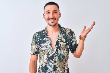 Young handsome man wearing Hawaiian summer shirt over isolated background smiling cheerful presenting and pointing with palm of hand looking at the camera.